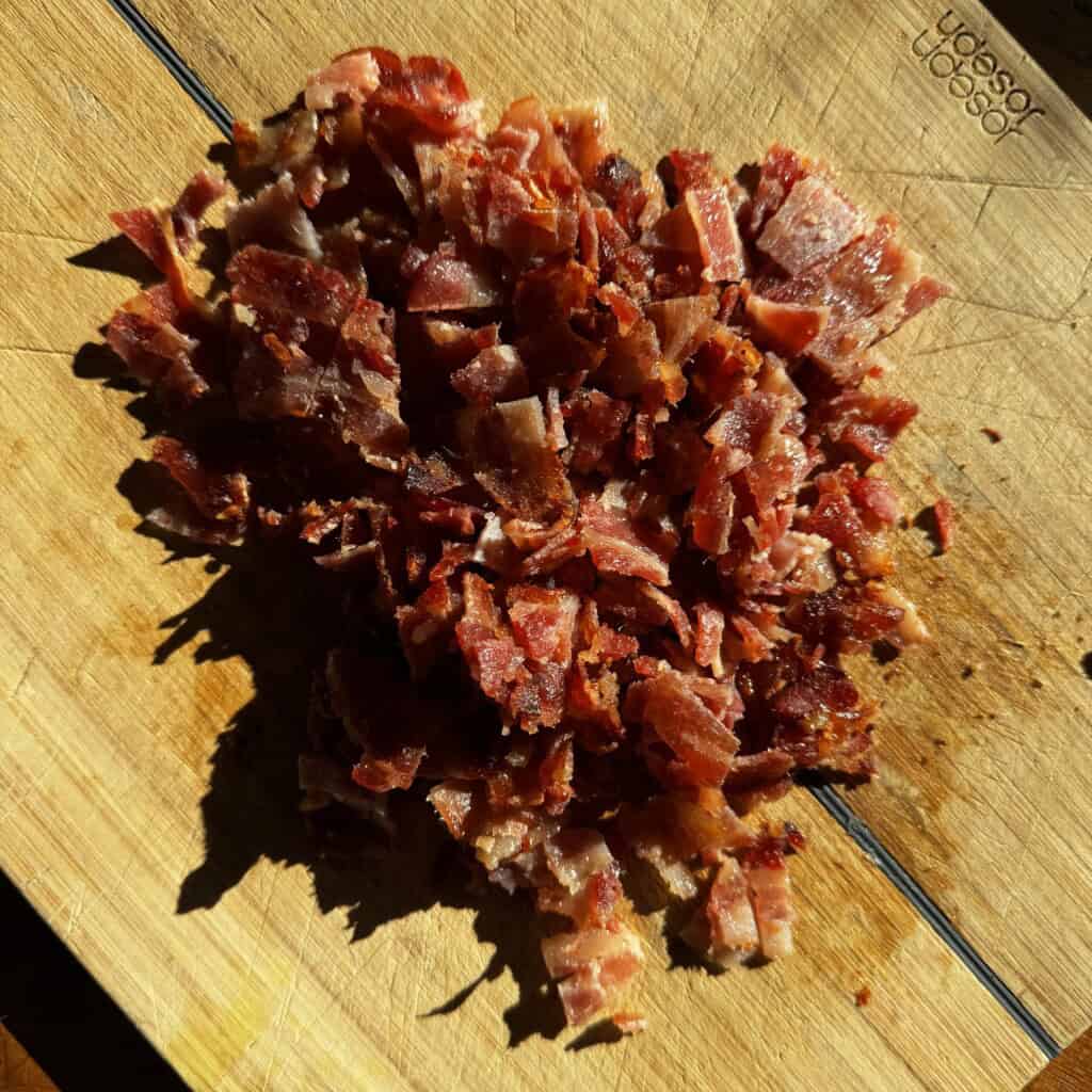 Chopped cooked bacon pieces are piled on a wooden cutting board in bright sunlight, showing crispy, reddish-brown texture.