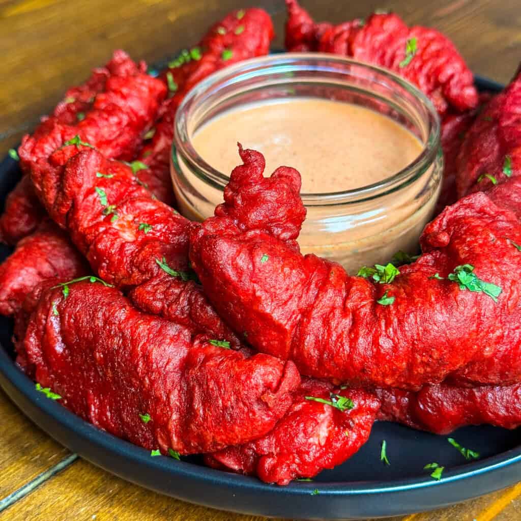 A plate of bright red fried chicken pakora garnished with chopped herbs, arranged in a circle around a glass bowl of creamy dipping sauce on a wooden table.