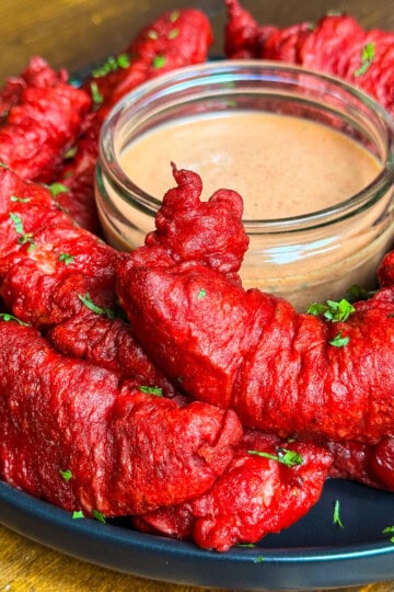 A plate of bright red fried chicken pakora garnished with chopped herbs, arranged in a circle around a glass bowl of creamy dipping sauce on a wooden table.