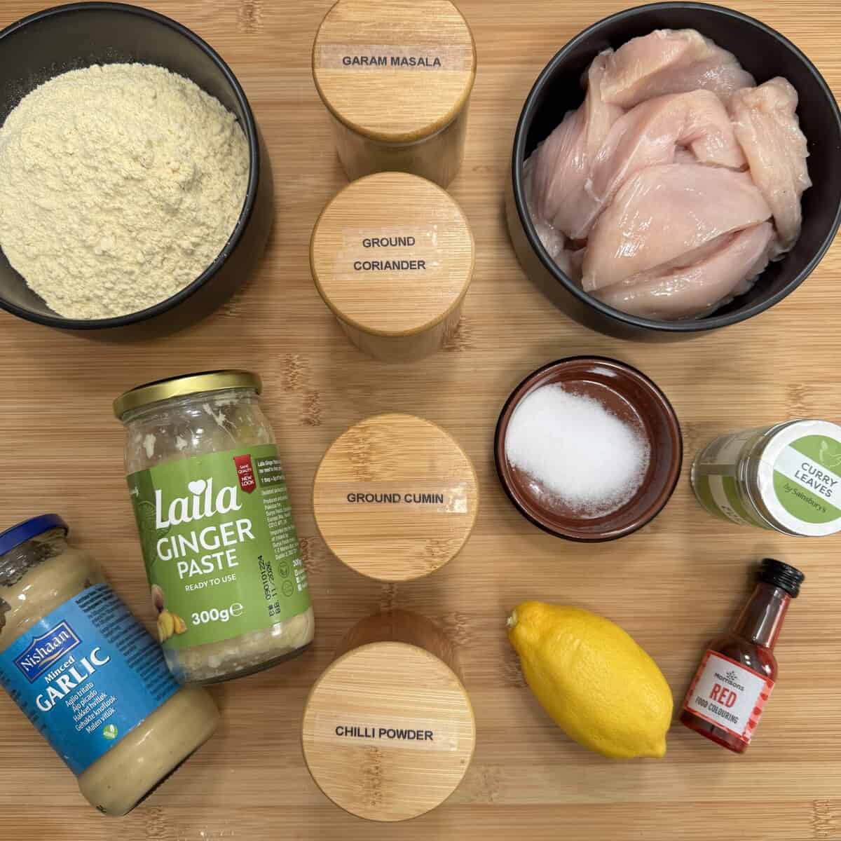 A top-down view of ingredients for chicken pakora: including raw chicken, gram flour, four spice jars (chilli, cumin, coriander and garam masala), ginger paste, garlic paste, curry leaves, salt, a lemon, and a bottle of red coloring, arranged on a wooden surface.