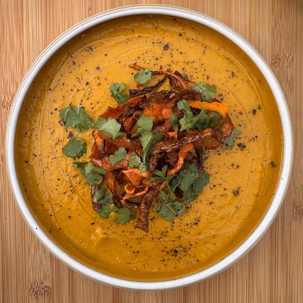 A bowl of creamy carrot and coriander soup, garnished with crispy vegetable strips, fresh coriander leaves, and cracked black pepper, sits on a wooden surface.