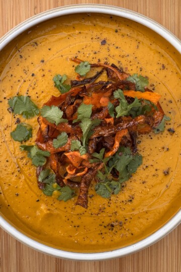 A bowl of creamy carrot and coriander soup, garnished with crispy vegetable strips, fresh coriander leaves, and cracked black pepper, sits on a wooden surface.