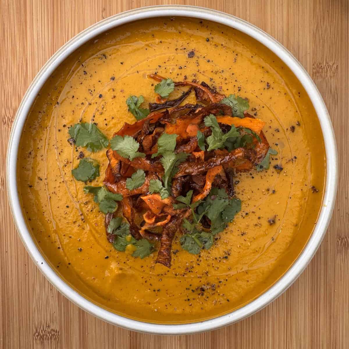 A bowl of creamy carrot and coriander soup, garnished with crispy vegetable strips, fresh coriander leaves, and cracked black pepper, sits on a wooden surface.