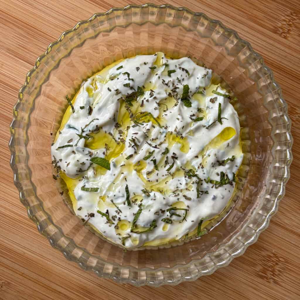 A glass bowl filled with creamy tzatziki, drizzled with olive oil and sprinkled with chopped fresh herbs, sits on a wooden surface.