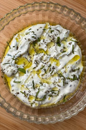 A glass bowl filled with creamy tzatziki, drizzled with olive oil and sprinkled with chopped fresh herbs, sits on a wooden surface.