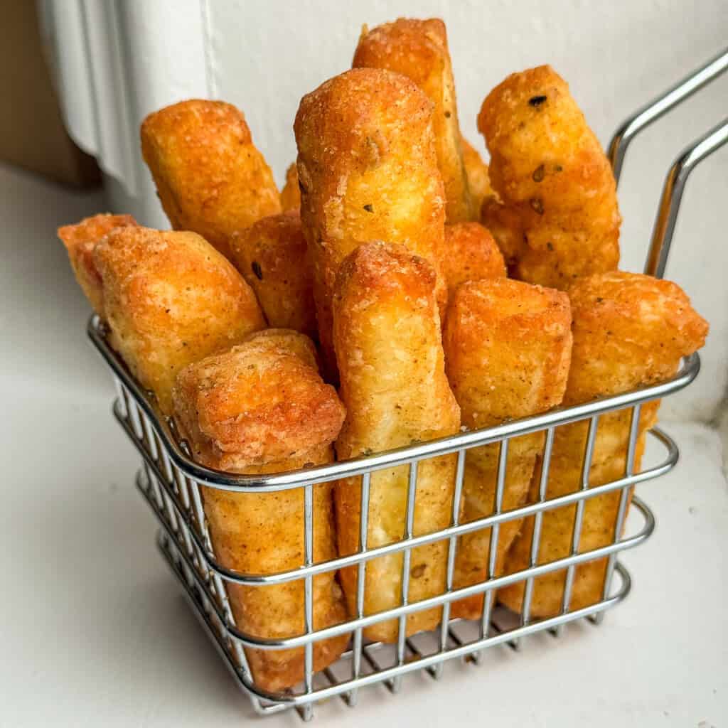 A metal basket filled with thick, golden-brown, crispy halloumi fries, placed on a white surface with a soft background.