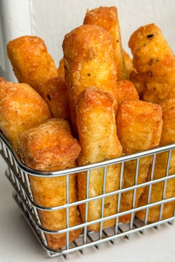 A metal basket filled with thick, golden-brown, crispy fries or breadsticks, placed on a white surface with a soft background.