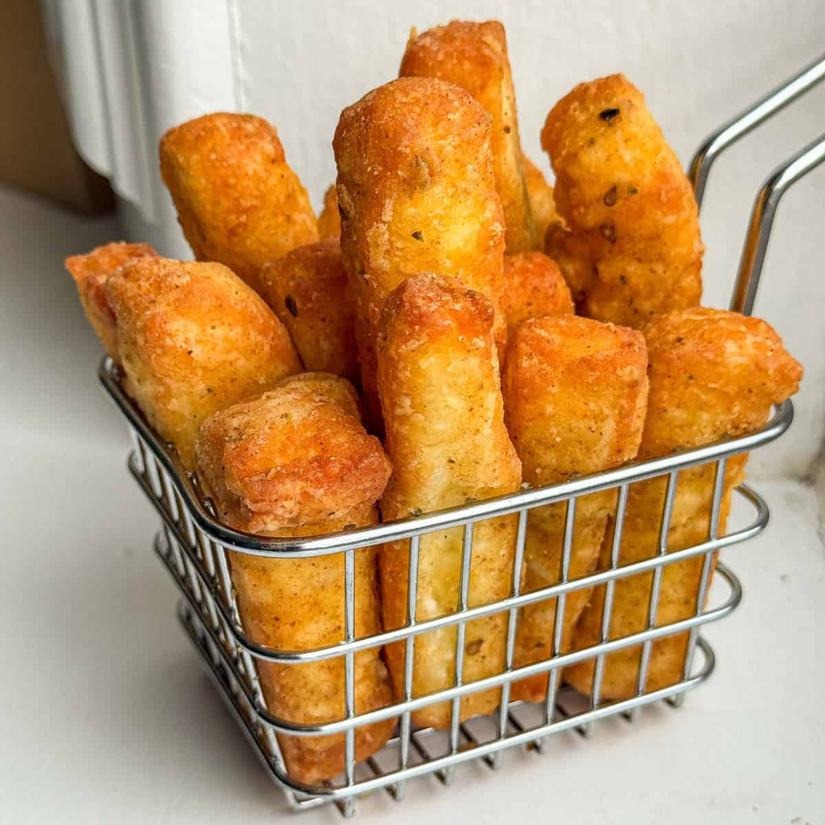 A metal basket filled with thick, golden-brown, crispy halloumi fries, placed on a white surface with a soft background.