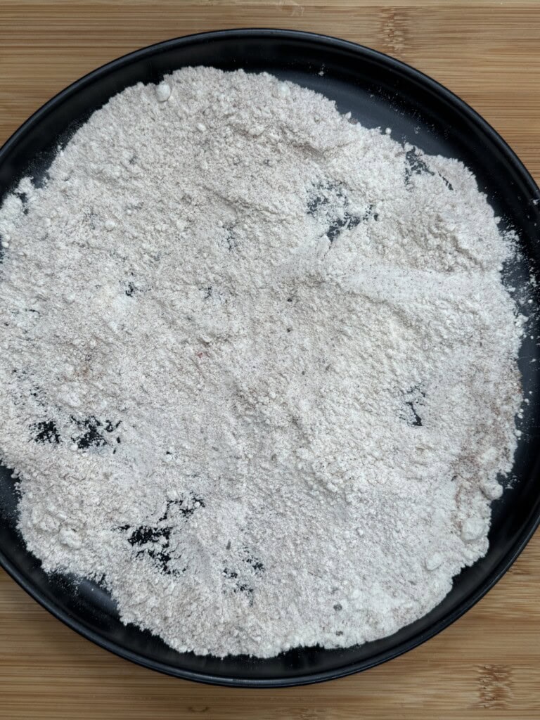 A black plate on a wooden surface filled with pieces of food coated in a light layer of flour, partially obscuring the items beneath the flour.