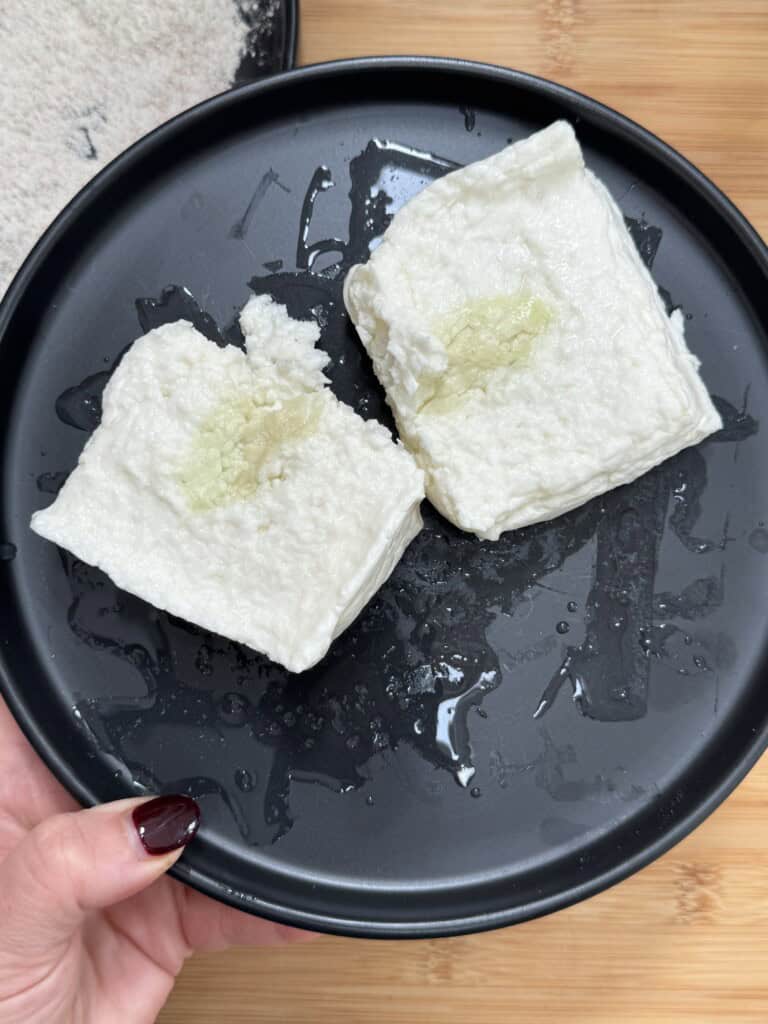 Two pieces of white tofu with grated ginger on top are placed on a black plate. A hand with dark red nail polish holds the plate. The plate has some water droplets. A wooden surface is visible underneath.