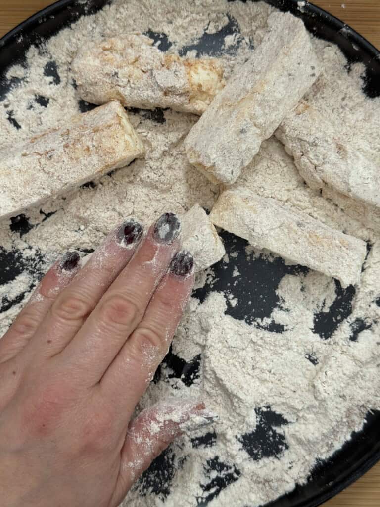 A hand with painted nails coats rectangular pieces of food in flour on a black plate, preparing them for cooking.