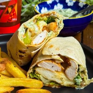 A close-up of a chicken wrap cut in half, showing grilled chicken, lettuce, and sauce inside a tortilla, served with golden fries on a black plate. A bowl of sauce and a bottle are in the background.