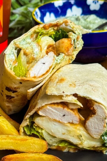 A close-up of a chicken wrap cut in half, showing grilled chicken, lettuce, and sauce inside a tortilla, served with golden fries on a black plate. A bowl of sauce and a bottle are in the background.