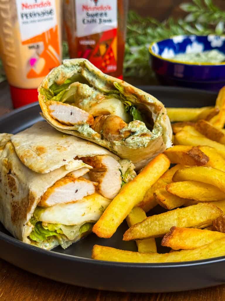 A plate with golden fries and a chicken wrap cut in half, showing grilled chicken, lettuce, and sauce inside. Bottles of sauces and a bowl are in the background.
