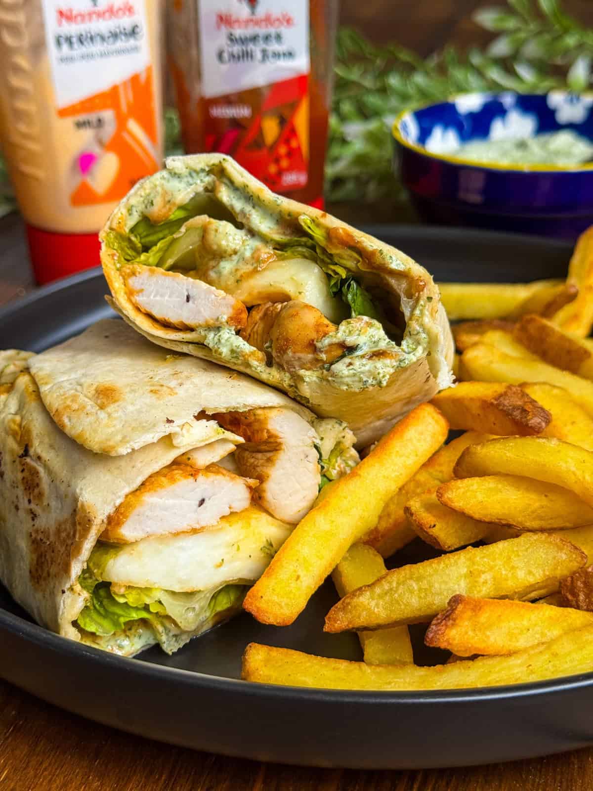 A plate with golden fries and a chicken wrap cut in half, showing grilled chicken, lettuce, and sauce inside. Bottles of sauces and a bowl are in the background.