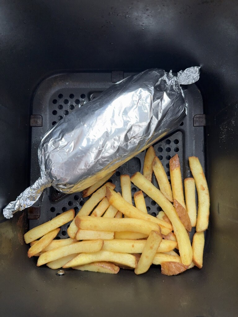 A foil-wrapped item and cooked French fries sit inside an air fryer basket. The fries are golden and scattered at the bottom, with the foil-wrapped food placed on a raised section above them.