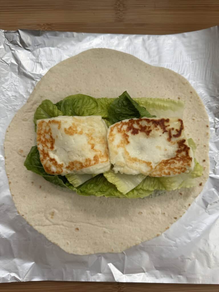 A tortilla wrap on foil topped with fresh green lettuce leaves and two browned slices of halloumi cheese.
