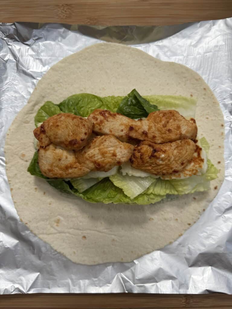 A tortilla wrap on foil, topped with green lettuce leaves and several pieces of grilled chicken, ready to be folded.