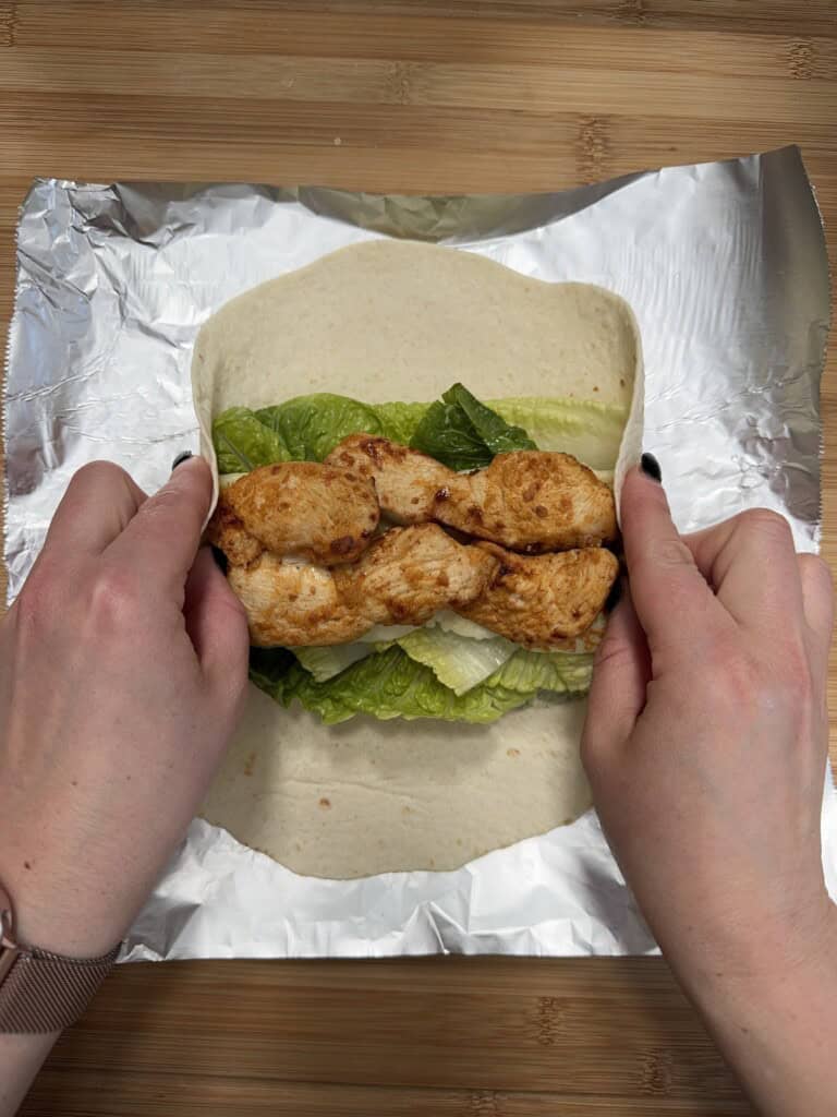 Two hands are folding a tortilla wrap with grilled chicken and lettuce, placed on a sheet of aluminum foil on a wooden surface.
