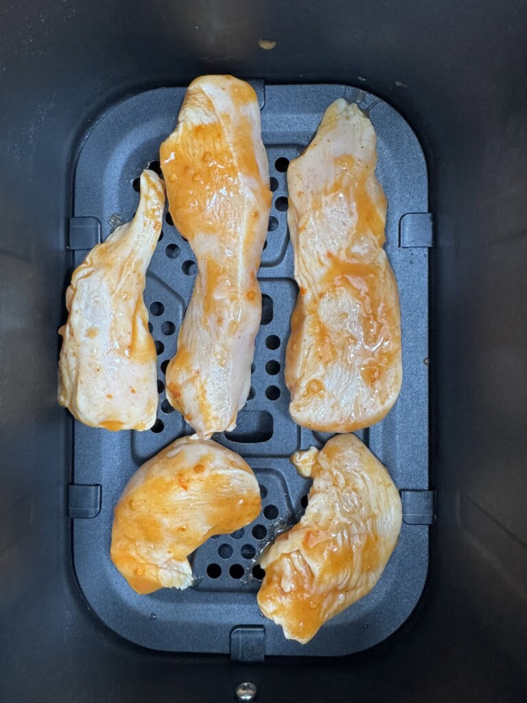 Five pieces of raw chicken coated with orange marinade are arranged inside an air fryer basket, ready to be cooked.