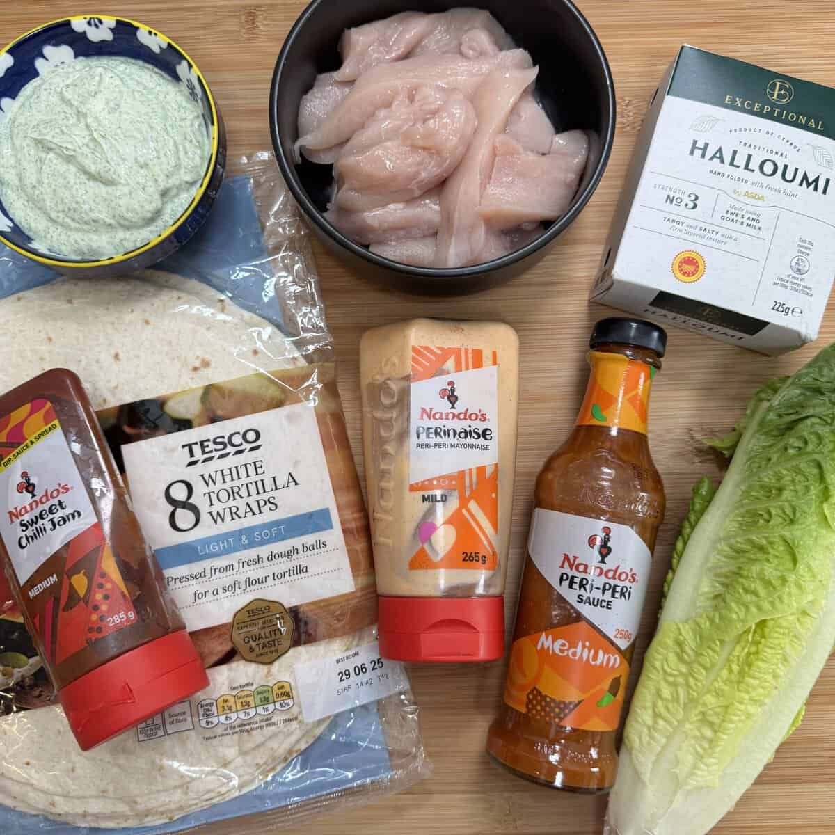 A wooden board displays chicken strips in a black bowl, a block of halloumi, romaine lettuce, white tortilla wraps, Nandos Perinaise, medium Peri-Peri sauce, sweet chilli jam, and a dish of green dip.