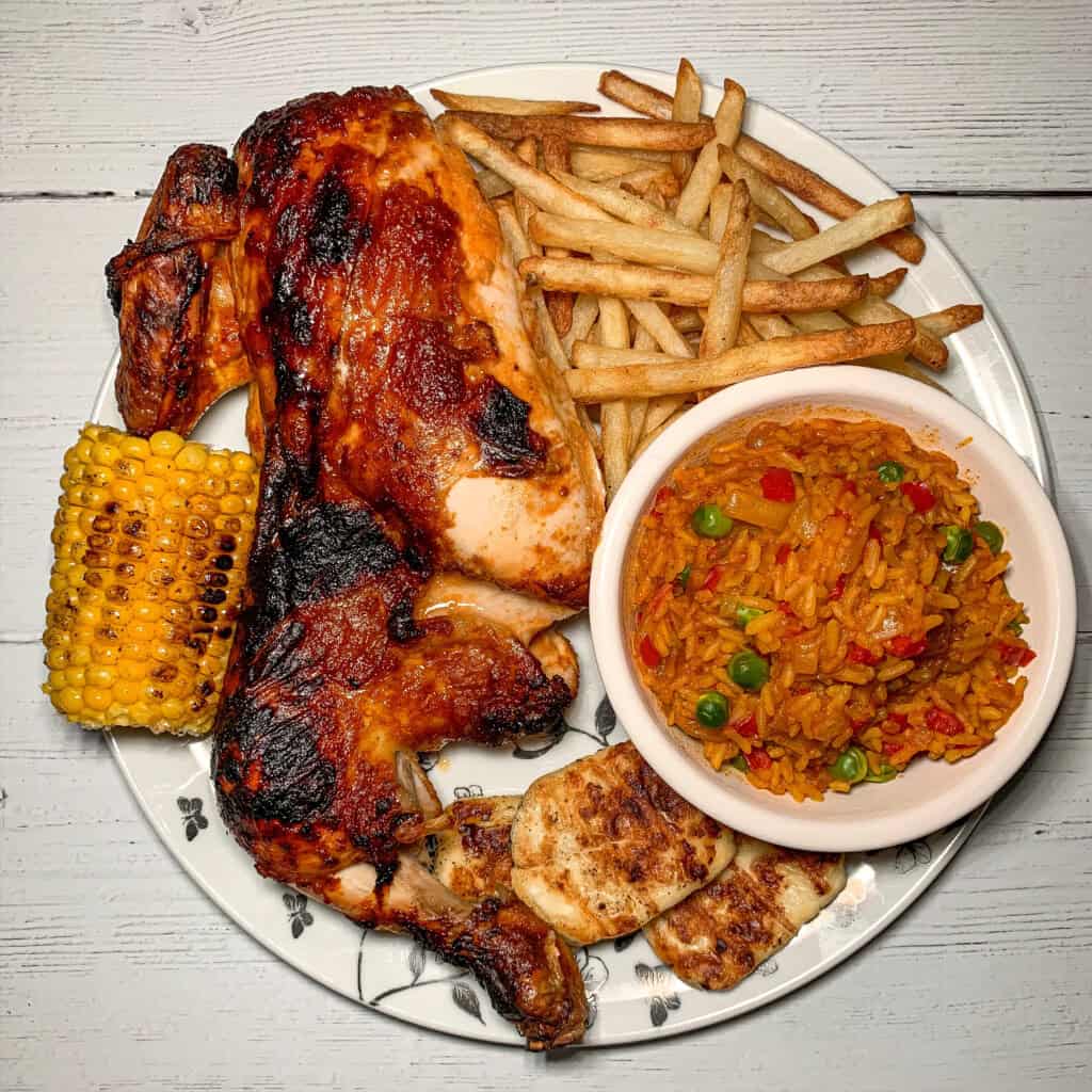 A plate with grilled chicken, French fries, a bowl of vegetable rice, grilled corn on the cob, and slices of grilled cheese, all served on a white plate with a floral pattern.