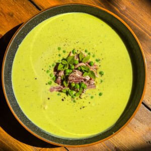 A bowl of creamy green pea soup garnished with shredded meat, green peas, fresh mint leaves, and a sprinkle of black pepper, served on a wooden table.
