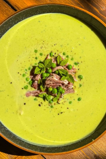 A bowl of creamy green pea soup garnished with shredded meat, green peas, fresh mint leaves, and a sprinkle of black pepper, served on a wooden table.