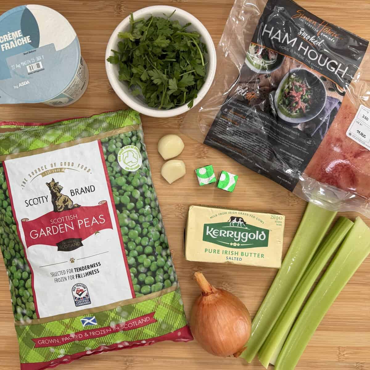 Ingredients for a soup recipe on a wooden surface: frozen peas, crème fraîche, fresh parsley, ham hough, garlic cloves, stock cubes, butter, celery sticks, and an onion.