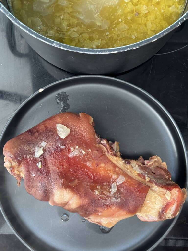A cooked pork knuckle with visible salt crystals rests on a black plate. Above it, a pot filled with broth and diced onions sits on a stovetop.