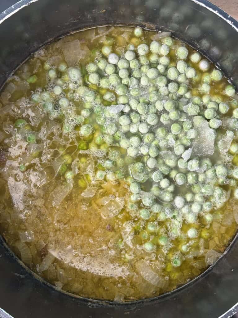 A close-up of a pot filled with soup containing diced onions and frozen peas in a yellowish broth. The peas are mostly grouped together and retain their frosty appearance.