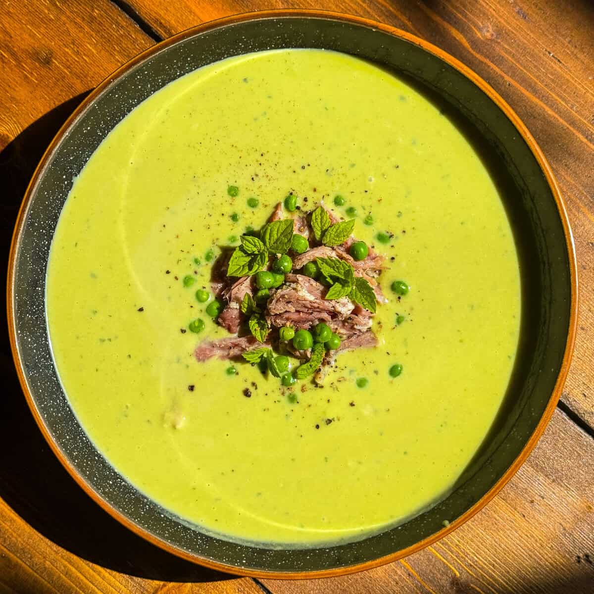 A bowl of creamy green pea and ham soup garnished with shredded ham, green peas, fresh mint leaves, and a sprinkle of black pepper, served on a wooden table.