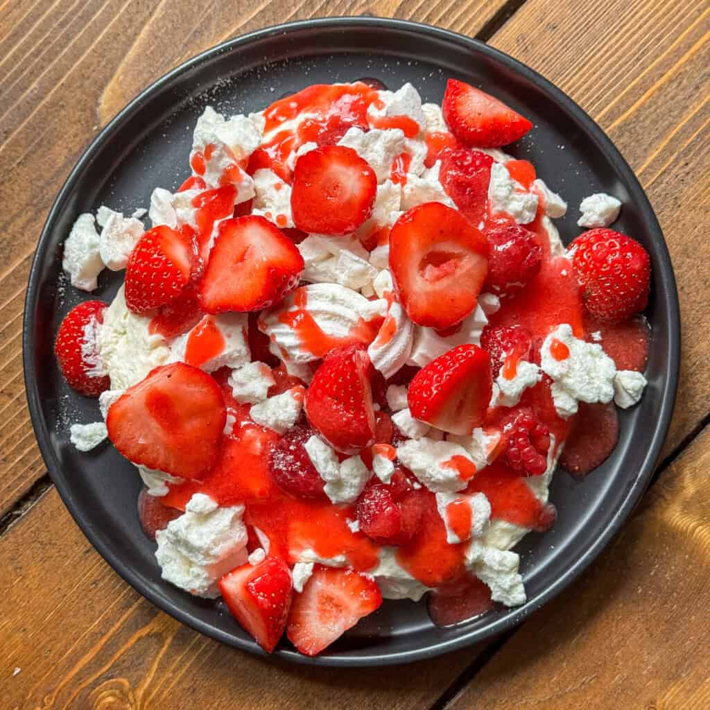 A black plate filled with Eton mess - meringue pieces, sliced fresh strawberries, and strawberry sauce, all arranged on a wooden table.