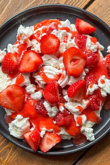 A black plate filled with Eton mess - meringue pieces, sliced fresh strawberries, and strawberry sauce, all arranged on a wooden table.