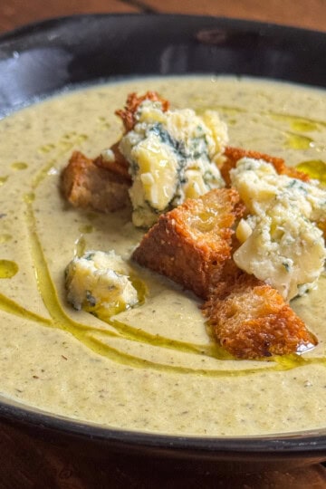 A bowl of creamy broccoli and blue cheese soup topped with golden croutons, chunks of blue cheese, and a drizzle of olive oil, served on a dark plate over a wooden table.