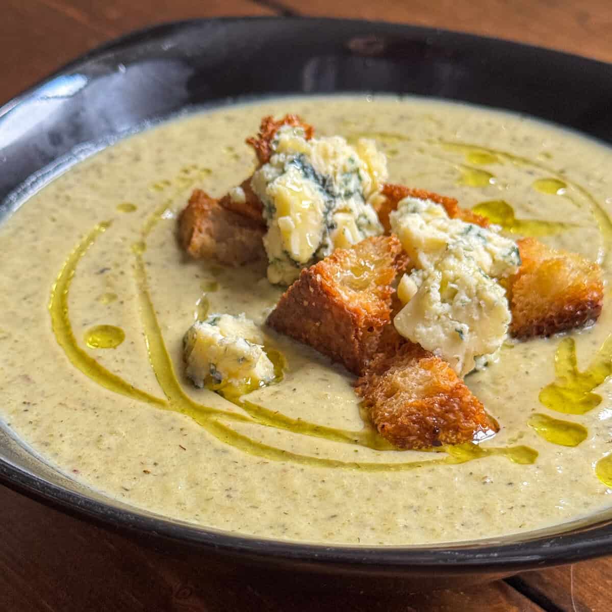 A bowl of creamy broccoli and blue cheese soup topped with golden croutons, chunks of blue cheese, and a drizzle of olive oil, served on a dark plate over a wooden table.