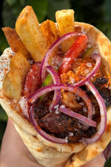 A hand holds a chicken gyros wrap filled with grilled meat, French fries, sliced red onions, tomatoes, tzatziki sauce, and sprinkled spices, with a blurred green background.