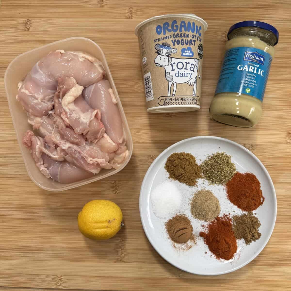 Raw chicken thighs, a tub of Greek-style yogurt, a jar of minced garlic, a lemon, and a white plate with various spices arranged in small piles, all on a wooden surface.