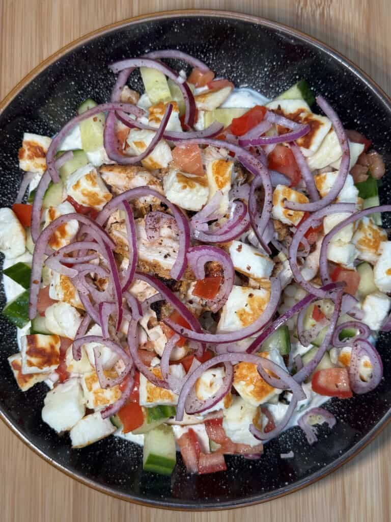 A bowl of salad with chopped cucumbers, tomatoes, grilled halloumi cheese, and thinly sliced red onions on a dark plate, placed on a wooden surface.