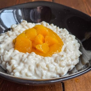 A black bowl filled with creamy rice pudding, topped with a generous portion of bright orange peach slices, sits on a wooden surface.