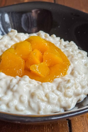 A black bowl filled with creamy rice pudding, topped with a generous portion of bright orange peach slices, sits on a wooden surface.
