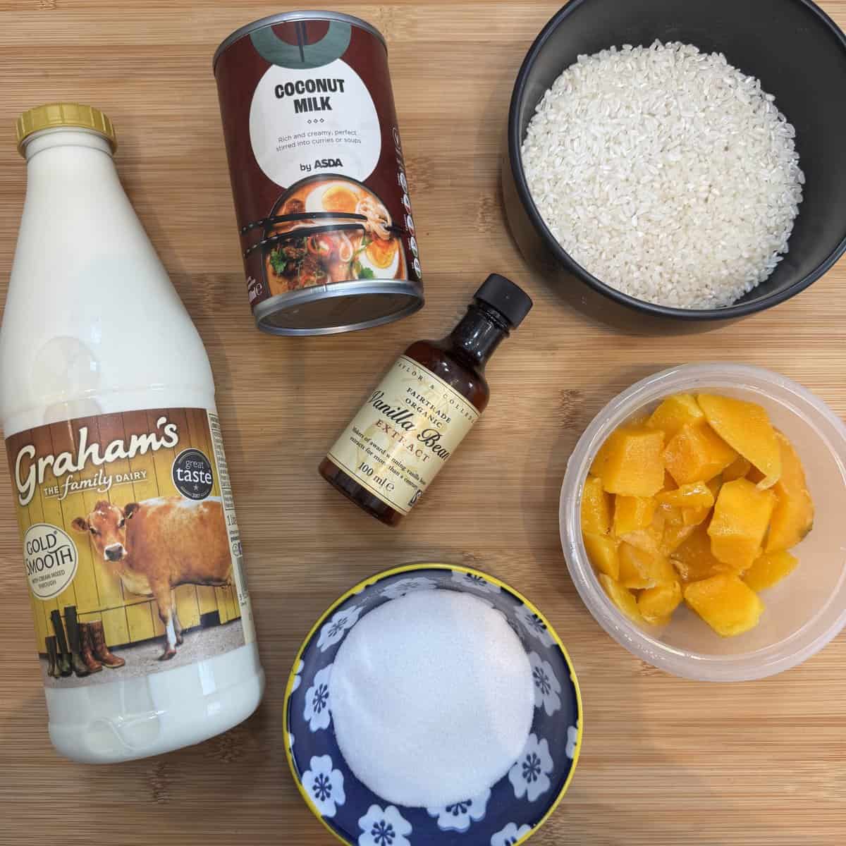 Ingredients for coconut rice pudding: bottle of milk, a can of coconut milk, a bowl of rice, a container of chopped mango, a bowl of sugar, and a bottle of vanilla extract arranged on a wooden surface.