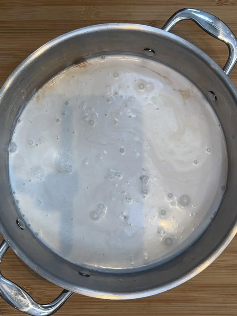 A stainless steel pot filled with a milky white liquid sits on a wooden surface. The liquid appears to have subtle swirls and small bubbles on its surface.