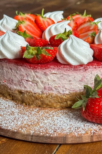 A Eton mess cheesecake with a crumbly base, topped with fresh strawberries and dollops of whipped cream, sits on a wooden board dusted with powdered sugar.