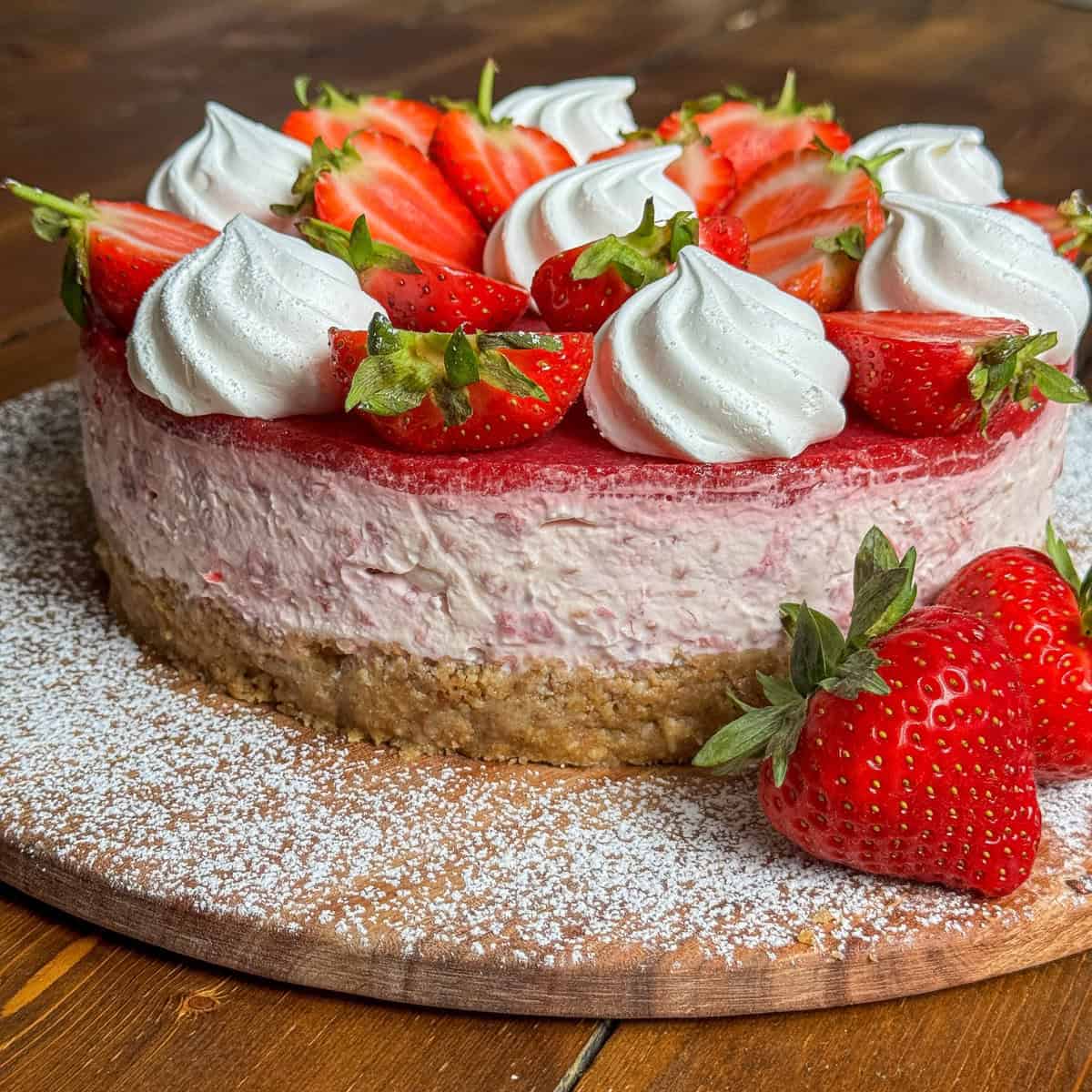 A Eton mess cheesecake with a crumbly base, topped with fresh strawberries and dollops of whipped cream, sits on a wooden board dusted with powdered sugar.