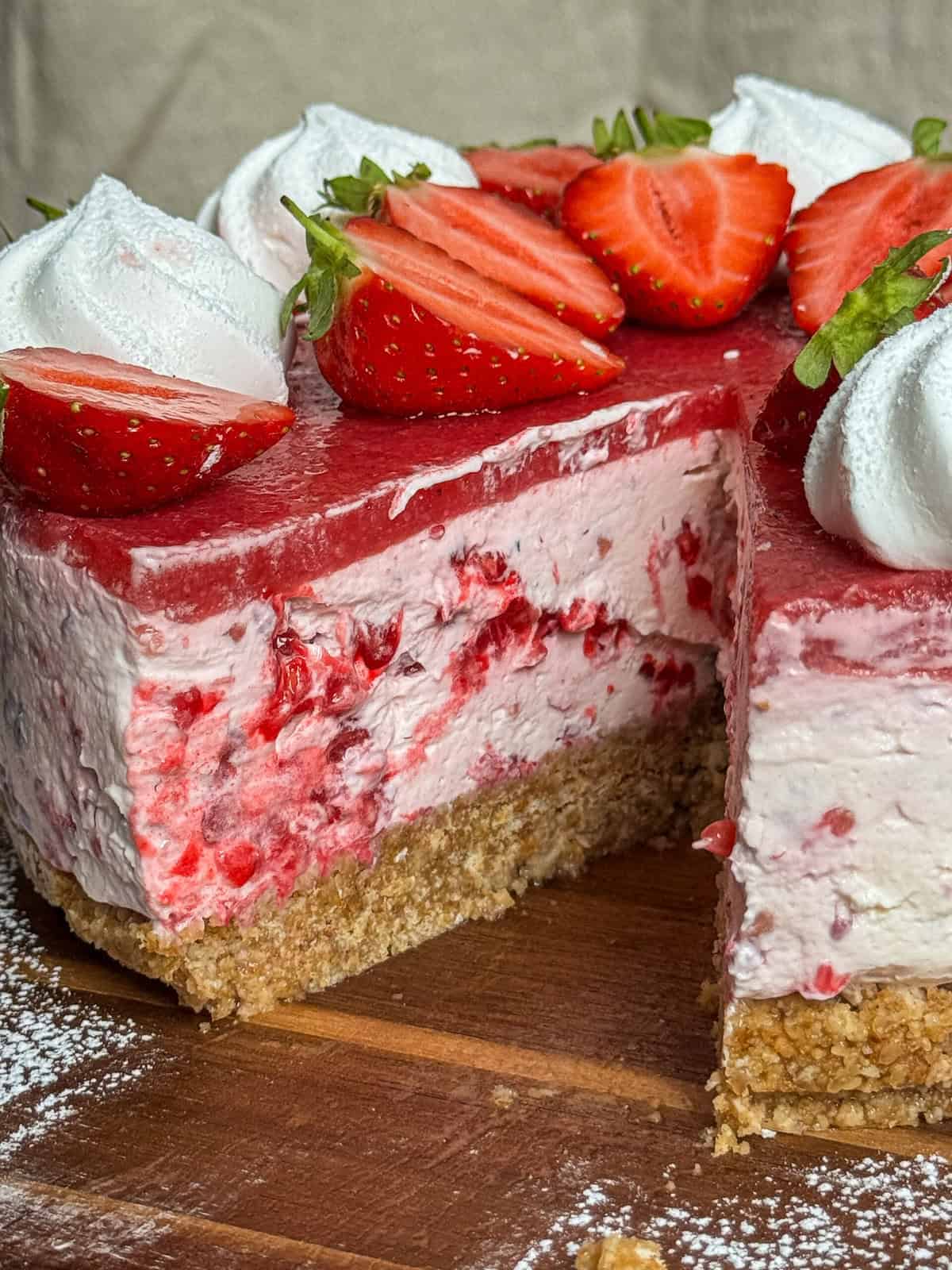A close-up of a sliced Eton mess cheesecake with a crumbly crust, creamy pink filling with berry pieces, a strawberry glaze, and fresh strawberry halves and whipped cream on top, displayed on a wooden board.