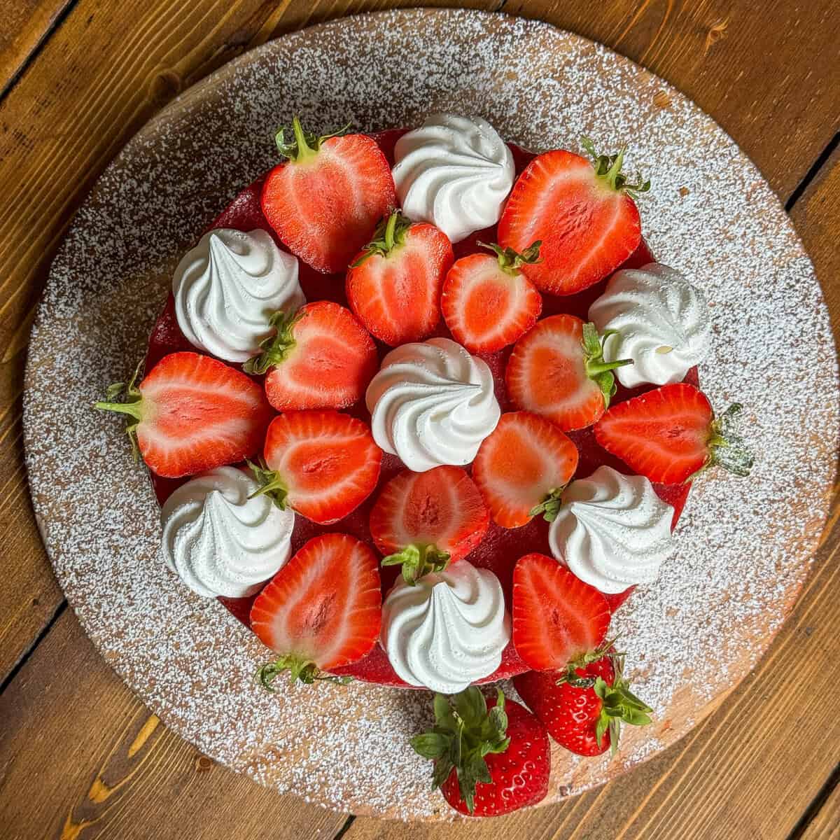 A round cake topped with halved strawberries and swirls of whipped cream, placed on a wooden platter sprinkled with powdered sugar. Two whole strawberries rest at the edge of the platter.
