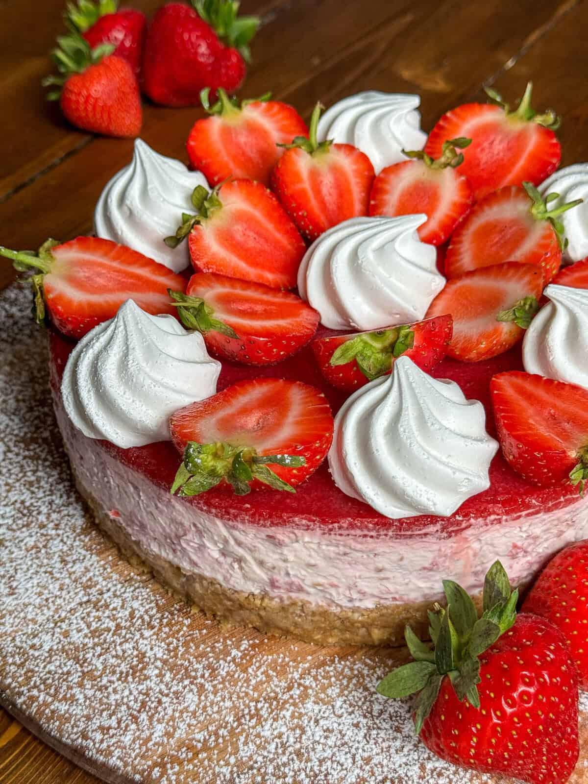 A Eton mess cheesecake topped with halved strawberries and swirls of whipped cream, set on a dusting of powdered sugar on a wooden board, with extra strawberries in the background.