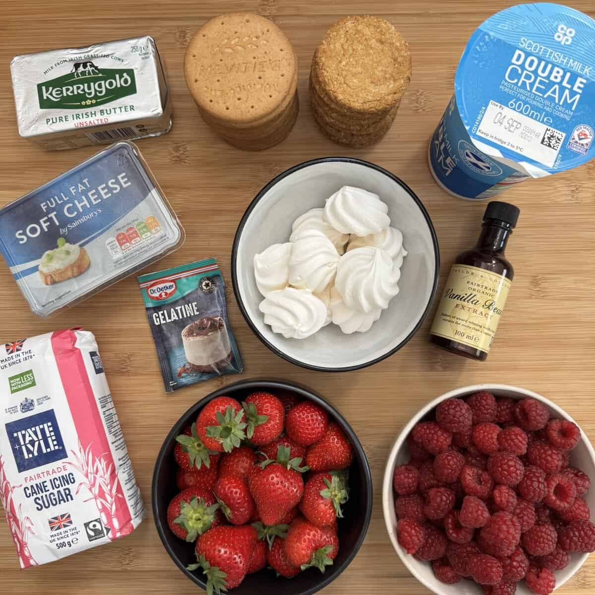 A top-down view of ingredients on a wooden surface: butter, digestive biscuits, oat biscuits, double cream, soft cheese, gelatine, vanilla extract, icing sugar, strawberries, raspberries, and a bowl of meringues.