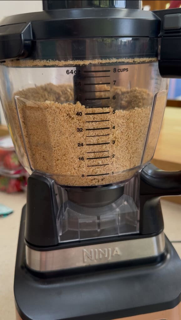 A food processor labeled NINJA filled with finely ground brown crumbs, showing measurement markings up to 8 cups on a clear container. The appliance sits on a kitchen counter.